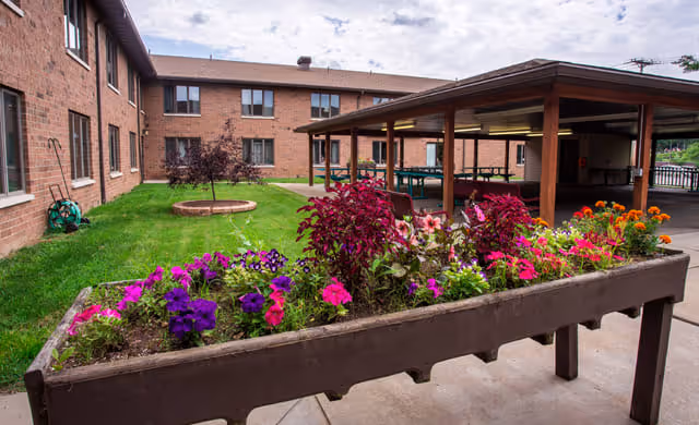 Outdoor courtyard area at a senior living facility with a raised flower bed filled with colorful flowers in the foreground, a grassy area with a circular garden bed, and a covered seating area. The building is a two-story brick structure with multiple windows.