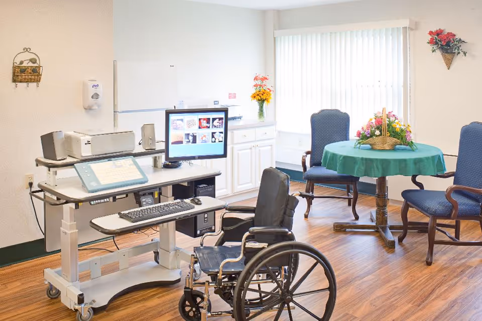 A bright room with a computer workstation on a mobile desk, a wheelchair in front of it, and a round table with a green tablecloth and a basket of flowers. The room has wooden flooring, white walls, vertical blinds on the window, and two blue upholstered chairs around the table. There are also decorative flower arrangements on the walls and a cabinet with a vase of flowers.
