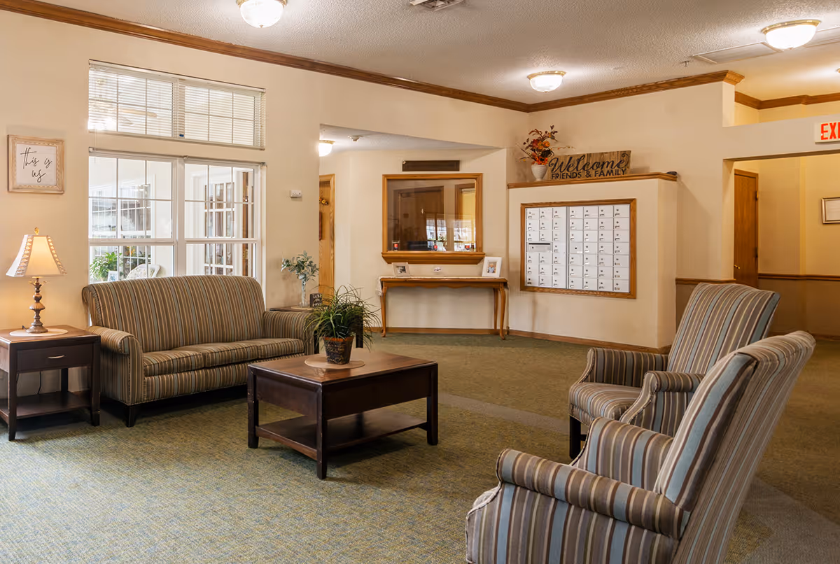 A cozy living room area in a senior living facility with striped upholstered chairs and a sofa, a wooden coffee table with a potted plant, a side table with a lamp, and a wall with mailboxes and a welcome sign. The room has beige walls, carpeted floors, and ceiling lights.