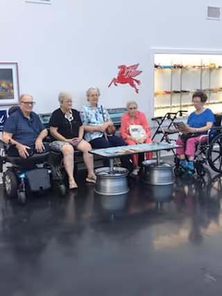Five elderly individuals sitting in a row inside a room with a polished dark floor. Two of them are in wheelchairs, and the others are seated on chairs. Behind them is a white wall with a red Pegasus logo and a display case with various items.