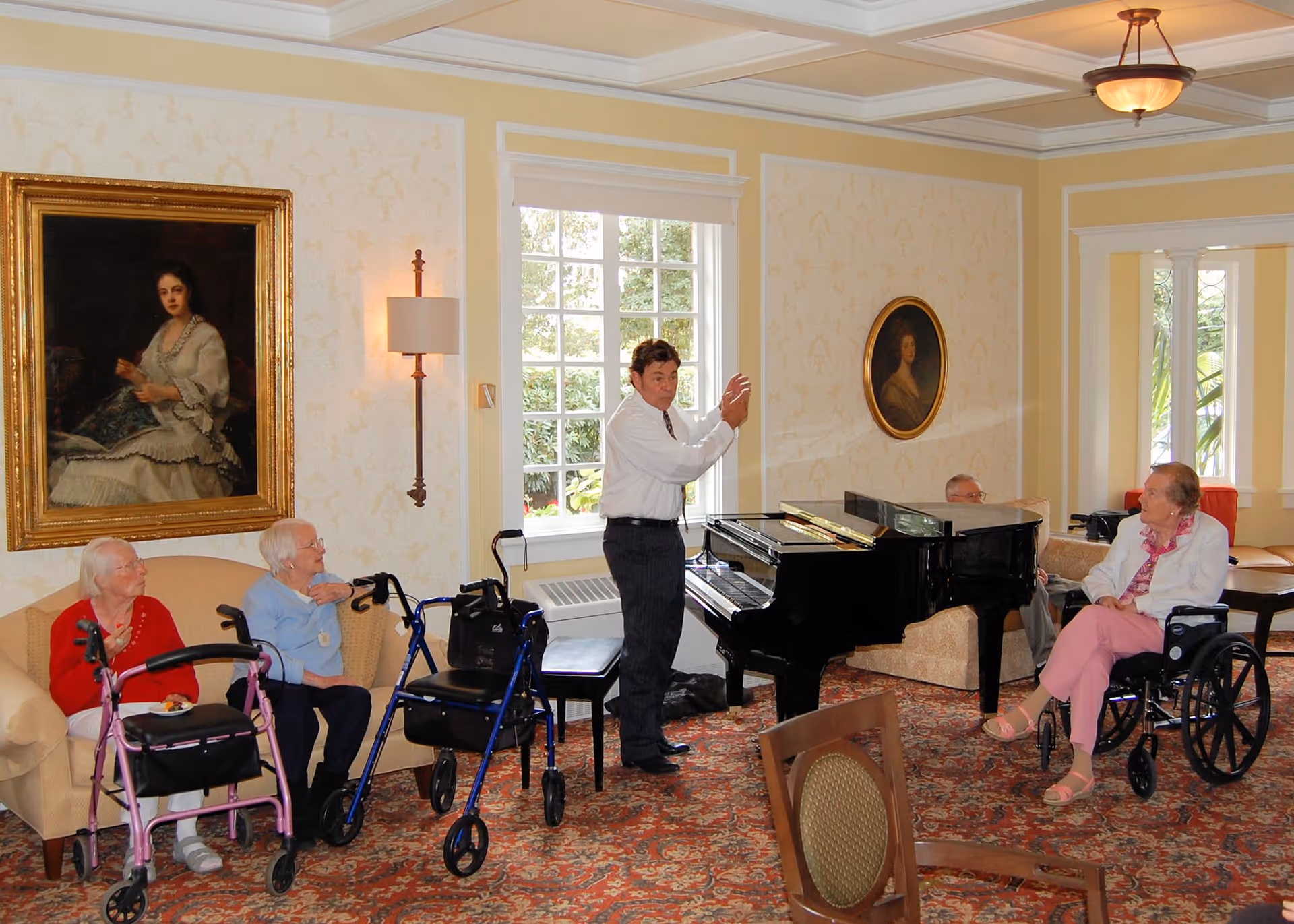 A group of elderly people sitting in a well-decorated living room with a man standing near a grand piano, seemingly performing or speaking to them. The room has patterned wallpaper, framed portraits, a large window, and a carpeted floor.