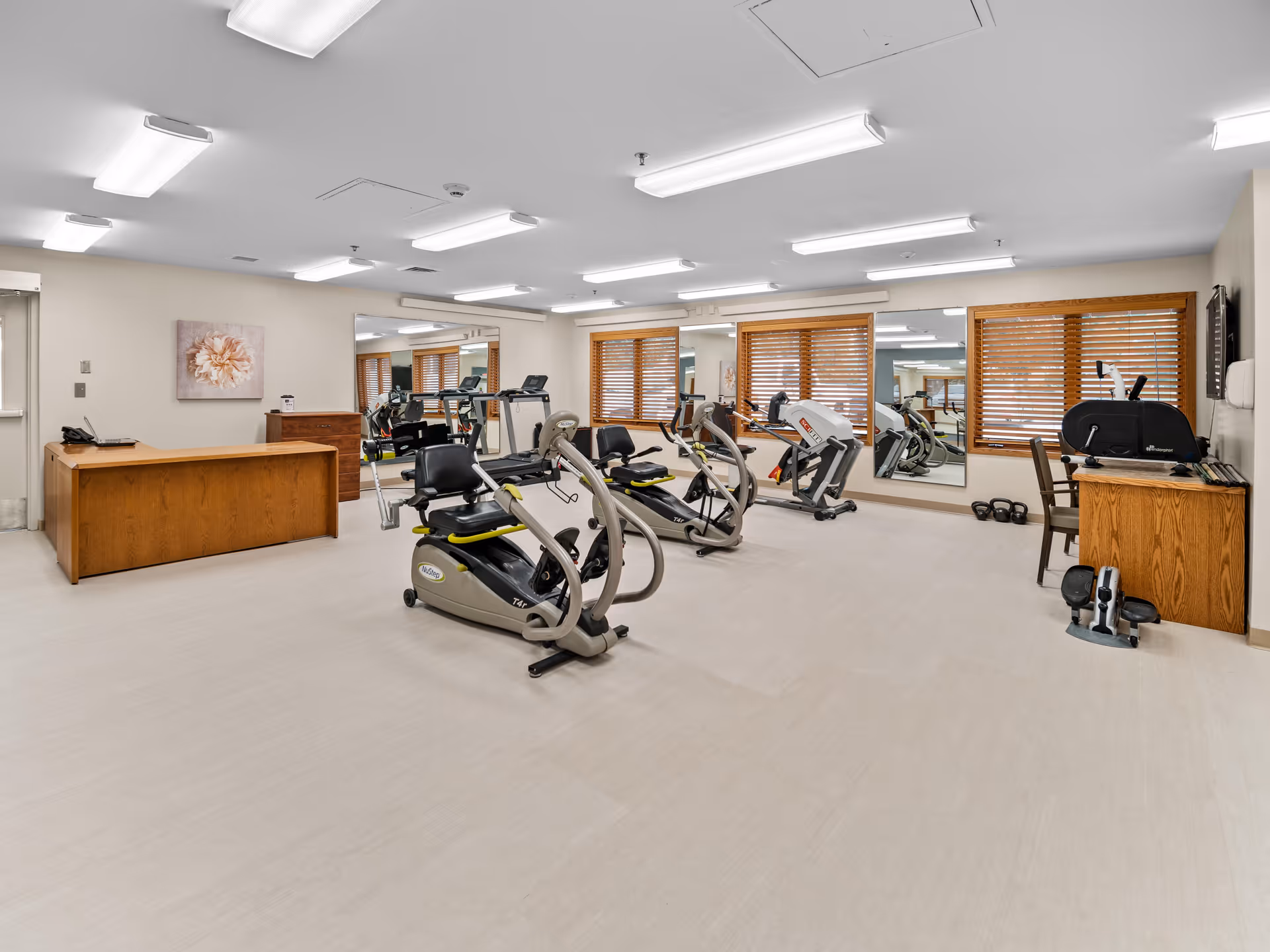 A well-lit exercise room with several pieces of cardio equipment including recumbent bikes and a treadmill. The room has large mirrors on the walls, wooden window blinds, and light-colored flooring. There is a wooden desk and chair in the corner, along with some small weights on the floor.
