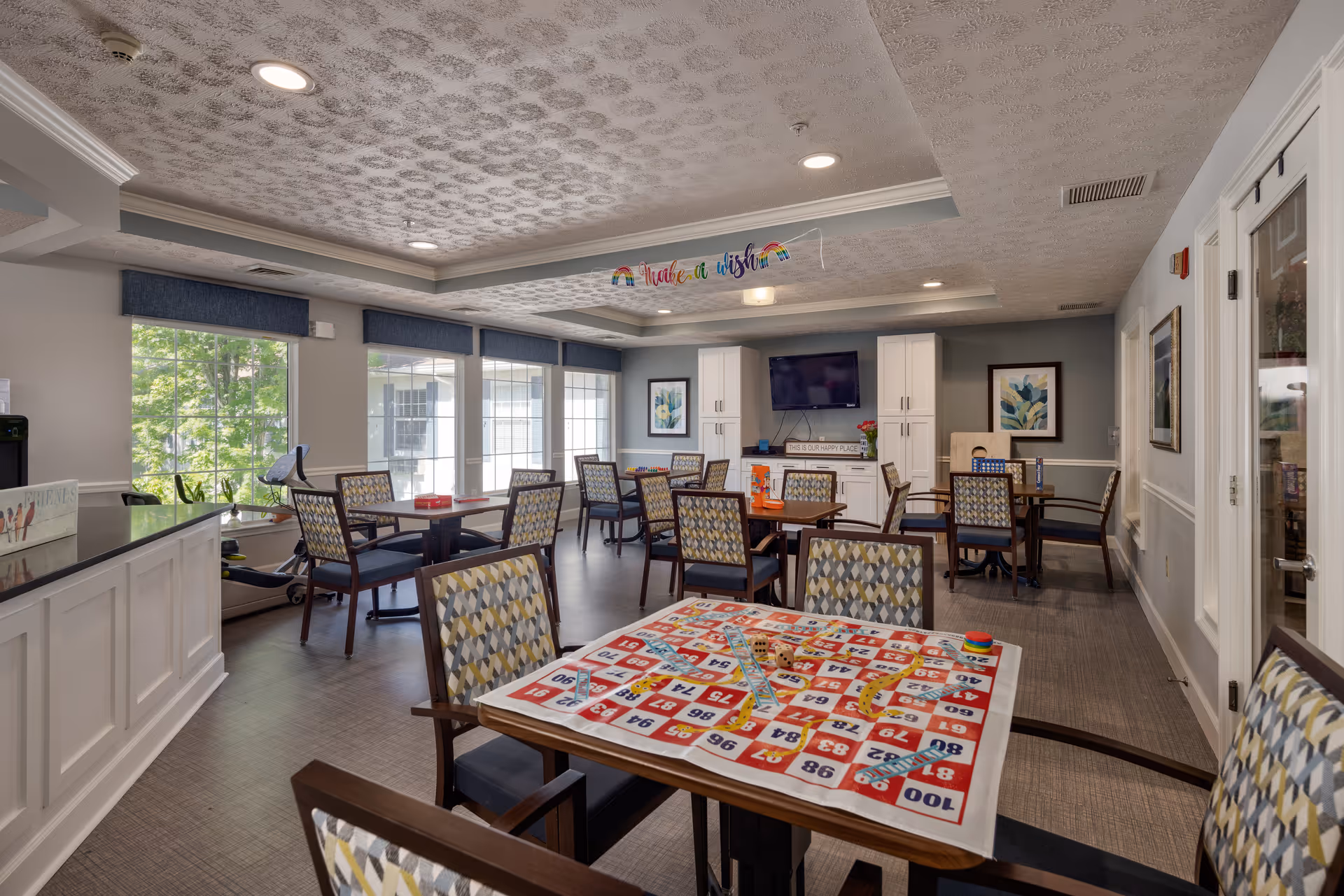 A bright and spacious common activity room with multiple tables and chairs arranged for group activities. One table in the foreground has a large board game set up. The room features large windows with blue valances letting in natural light, a wall-mounted TV, white cabinets, and framed artwork. A colorful banner hangs from the ceiling reading 'Make a wish'.