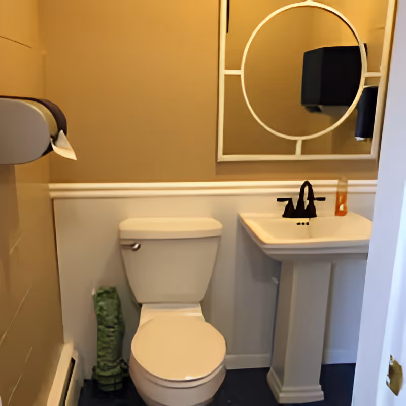 A small bathroom with a white toilet and a white pedestal sink. Above the sink is a round mirror with a decorative frame. There is a black faucet on the sink and a bottle of hand soap. A wall-mounted paper towel dispenser is visible on the left side. The walls are painted beige on the upper half and white on the lower half with a chair rail molding separating the two colors.