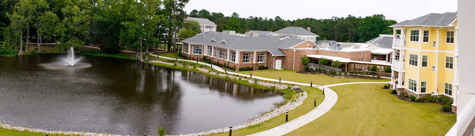 View of a senior living facility named Porters Neck Village featuring a large pond with a water fountain, surrounded by green trees and grass. A curved walking path runs alongside the pond, leading to multiple buildings including a yellow multi-story building and a brick building with large windows.