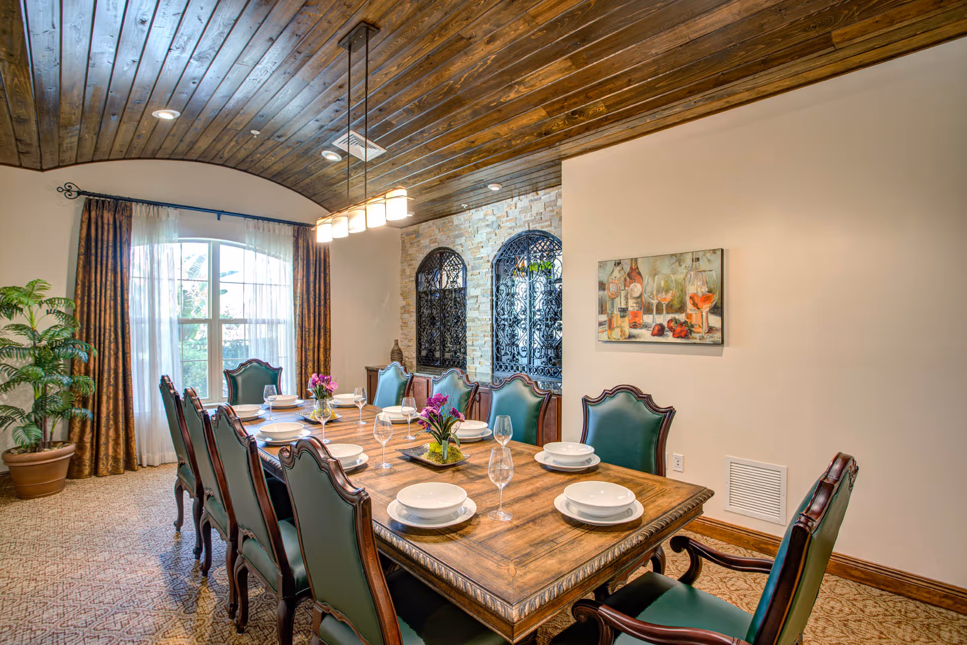 Formal dining room with a long wooden table set with plates and wine glasses, green upholstered chairs, wood-paneled ceiling and an arched window.