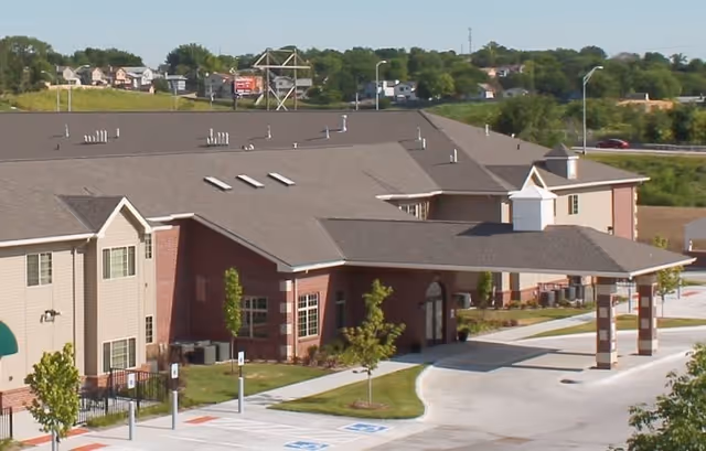 Exterior view of Richmont Village senior living facility showing a large building with a covered entrance, parking spaces including handicapped spots, and landscaped greenery around the building.