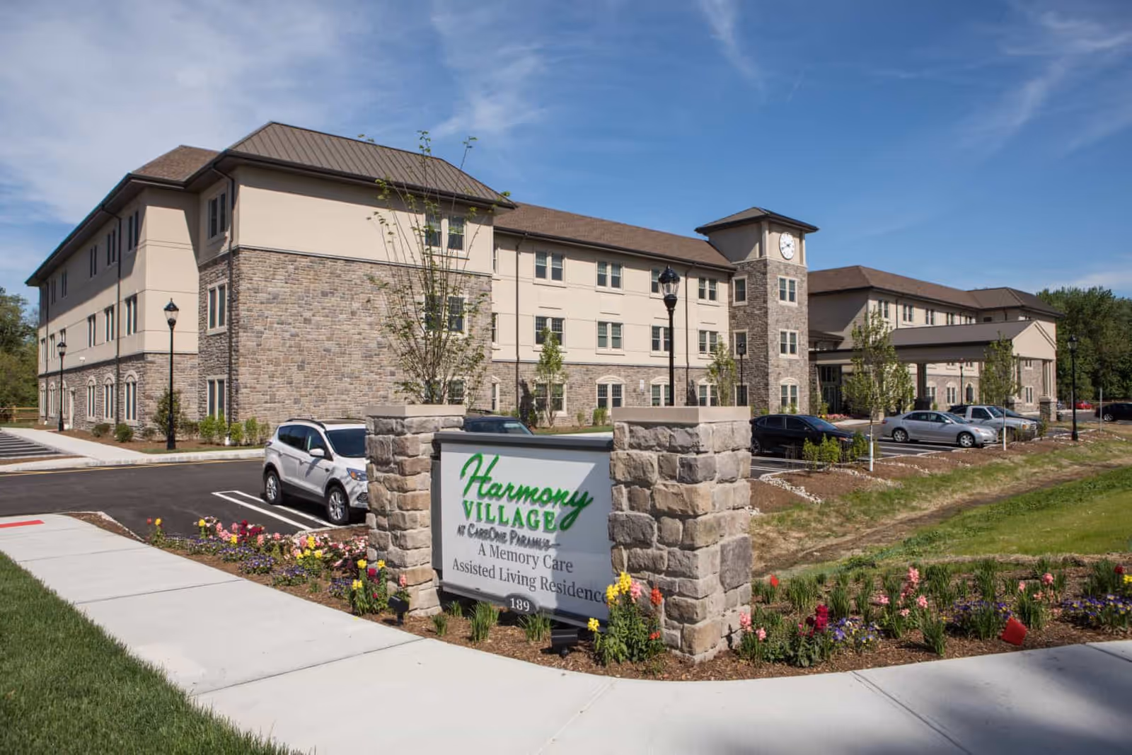 Exterior view of Harmony Village at CareOne Paramus, a three-story assisted living residence with stone and beige siding, a clock tower, landscaped flower beds, and a parking lot with several cars.