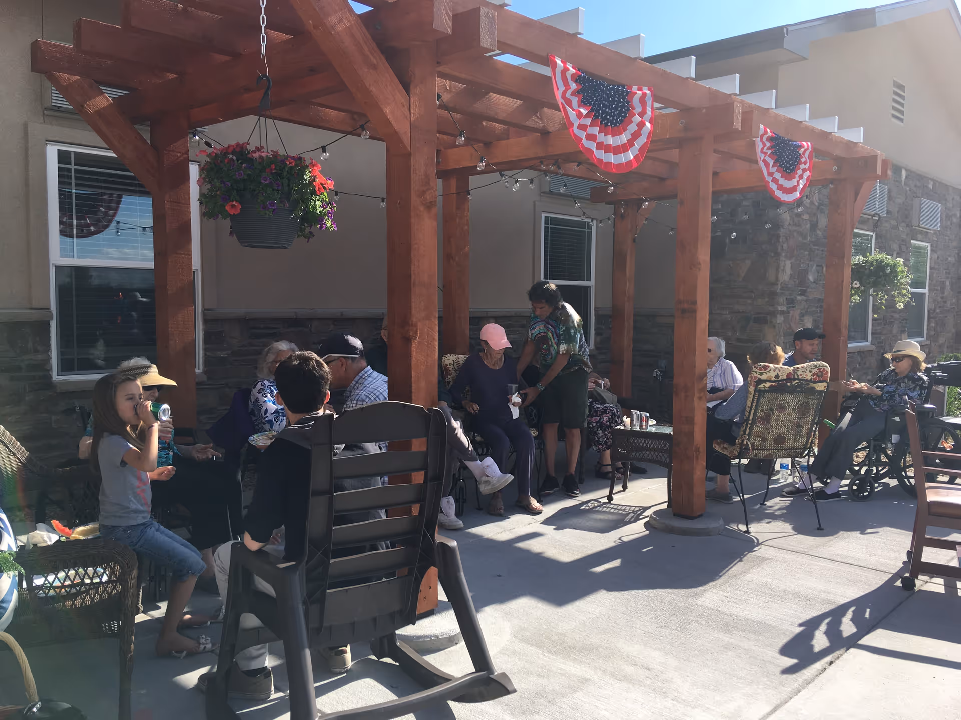 A group of seniors and visitors seated under a wooden pergola on an outdoor patio decorated with hanging flowers and American bunting.