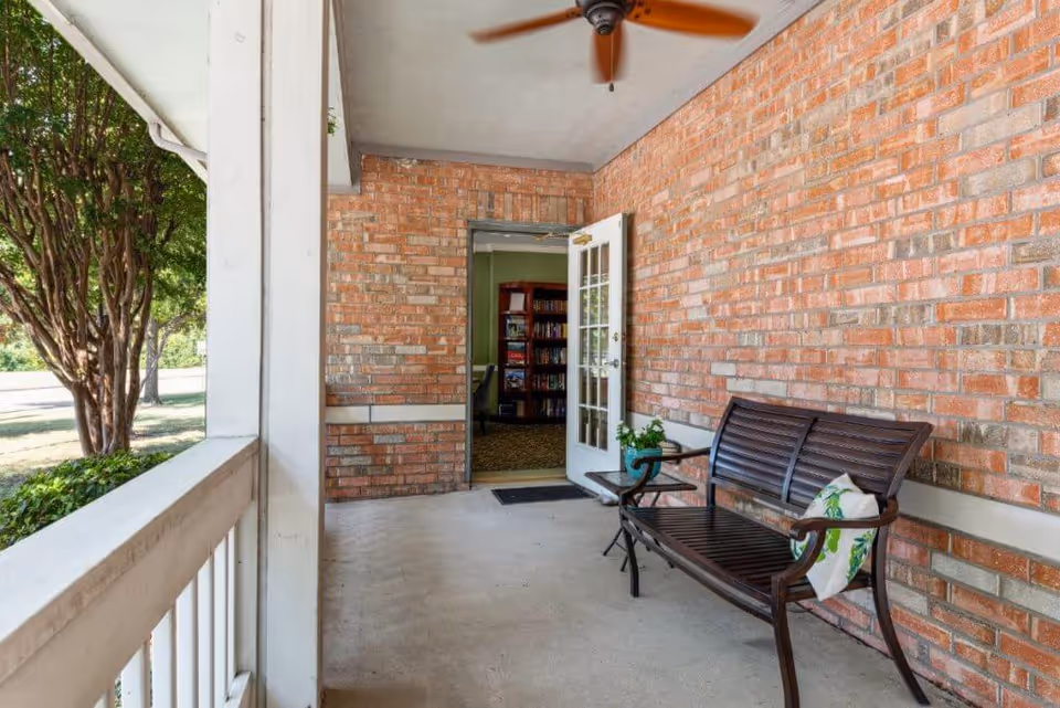 Covered porch area with a wooden bench that has a decorative pillow, a small side table with a potted plant, a ceiling fan above, and an open door leading inside to a room with bookshelves.