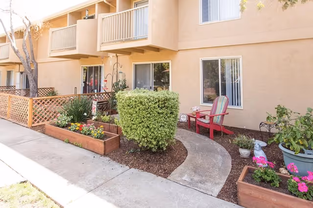 Outdoor garden area at Villa Serena Retirement Community featuring a curved concrete pathway, a trimmed bush, flower beds with colorful flowers, a red Adirondack chair with a small table, and potted plants near the building's beige exterior wall with windows and balconies above.