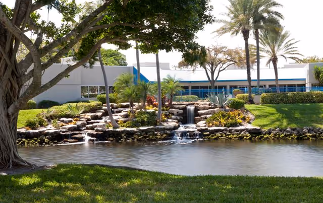 A landscaped outdoor area featuring a small pond with cascading waterfalls, surrounded by rocks, palm trees, and various plants. In the background, there is a modern building with large windows and a blue railing.