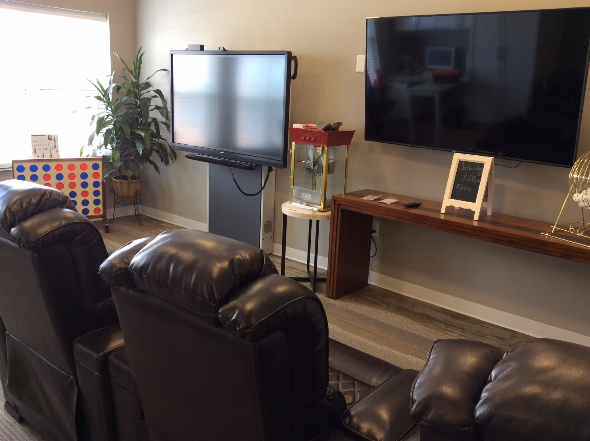 A cozy common area with three black leather recliners facing two large flat-screen TVs mounted on the wall. There is a small table with a popcorn machine between the TVs, a wooden console table beneath one TV with a small chalkboard sign, and a large Connect Four game and potted plant near a window with blinds.