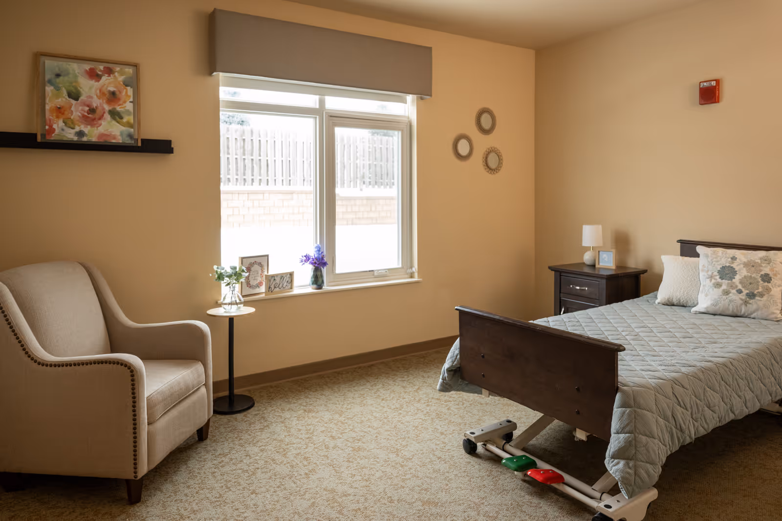 A cozy bedroom in a senior living facility with a single bed covered in a gray quilt, a beige armchair, a small round side table with a plant, and a window with a beige valance. The walls are painted light beige and decorated with a floral painting and three round mirrors. There is a dark wooden nightstand with a lamp and a small framed picture.