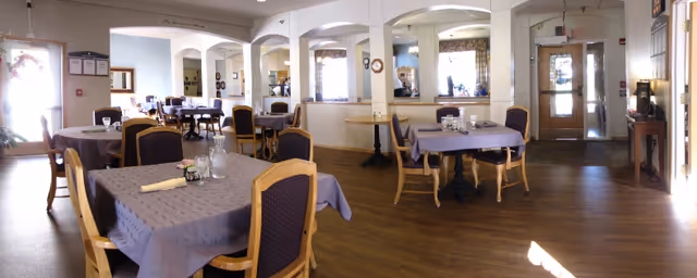 A dining room with several tables covered in gray tablecloths, each set with glasses, napkins, and small flower arrangements. Wooden chairs with dark cushions surround the tables. The room has wooden flooring, white walls with arches and mirrors, and windows with patterned curtains. There is a door at the far end and a small table with a lamp near the entrance.
