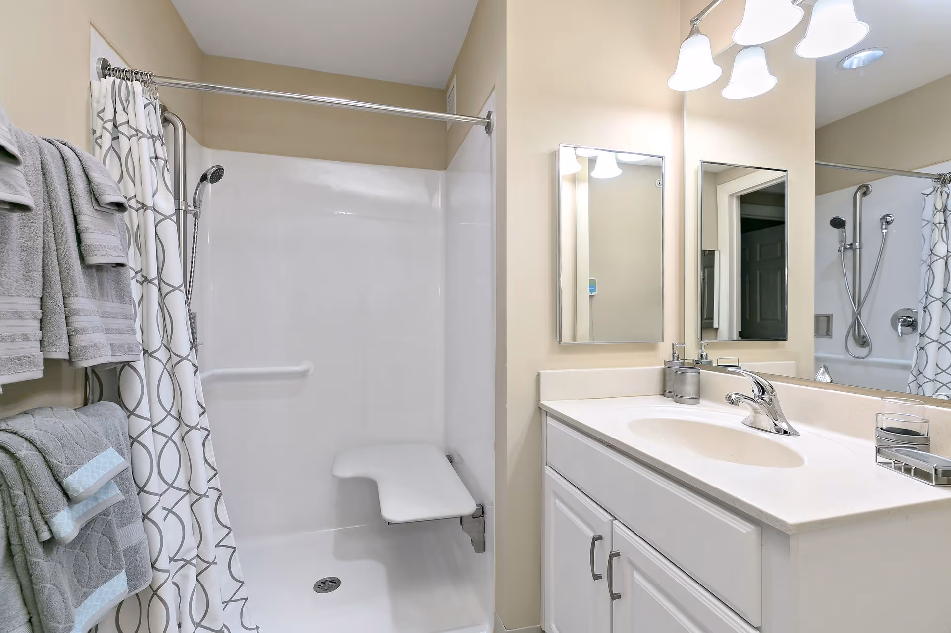 A clean bathroom featuring a white shower area with a foldable seat and a handheld showerhead. There is a patterned shower curtain and several gray towels hanging on a towel rack. The bathroom also has a white vanity with a sink, a faucet, soap dispensers, and two mirrors above the countertop. The walls are painted beige and the lighting fixture above the mirrors has three lights.