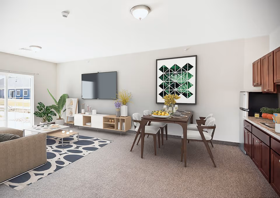A bright and modern living and dining area in a senior living facility. The room features a beige sofa, a coffee table on a patterned rug, a wall-mounted flat-screen TV, and a wooden dining table set with six chairs. There are decorative plants and vases with flowers, a large geometric green and black artwork on the wall, and a kitchenette with dark wood cabinets and a small refrigerator. Large sliding glass doors provide natural light and a view of the outdoor area.
