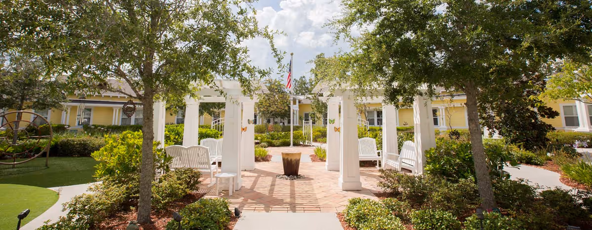Sunny courtyard with a white pergola and benches surrounded by trees and a yellow building in the background.