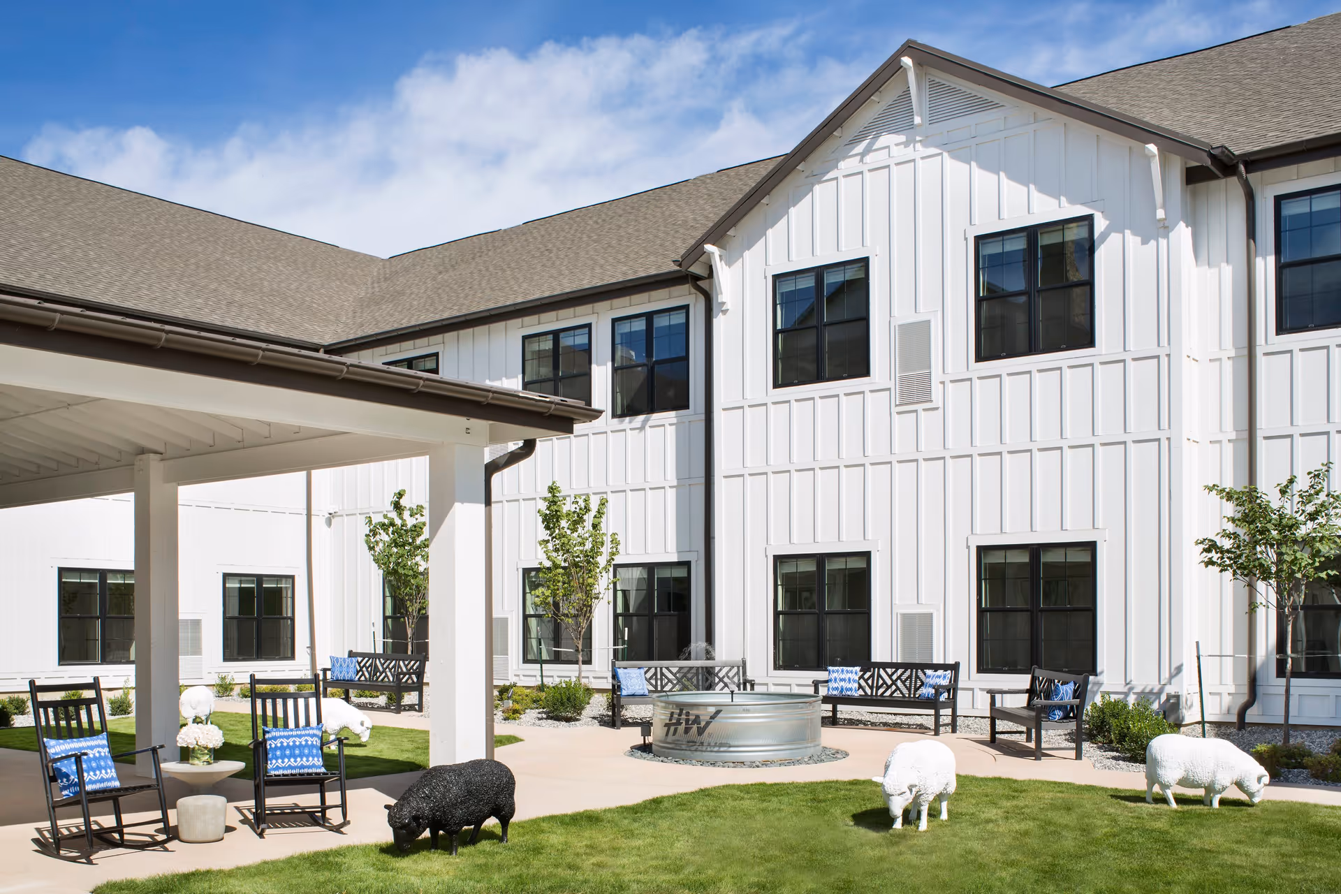 Outdoor courtyard area of a senior living facility with white two-story building in the background. The courtyard features green grass, several black benches with blue and white cushions, black rocking chairs, a covered patio area, and decorative sheep sculptures on the lawn. The sky is clear and blue.