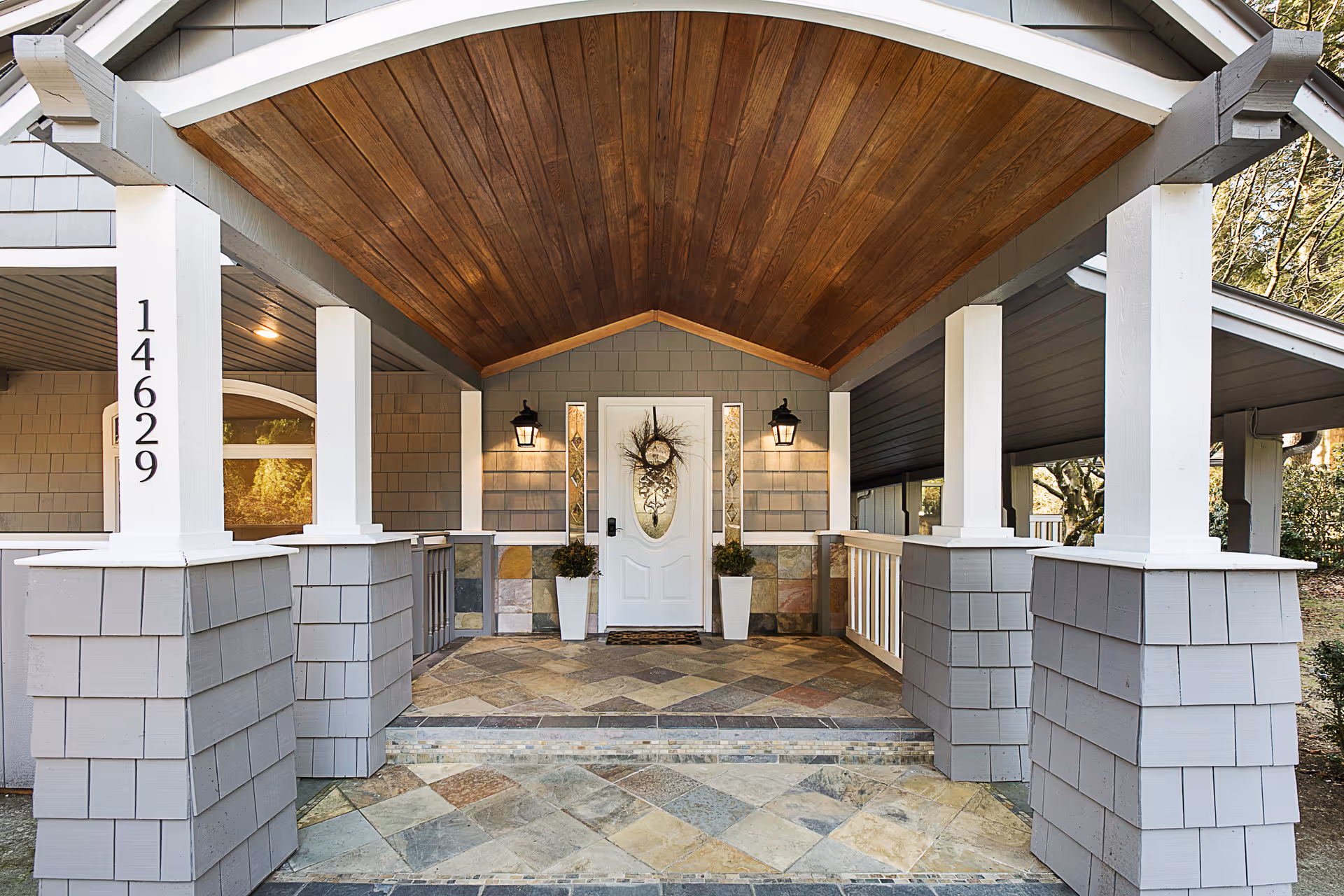 Covered front porch entrance of a house with a wooden ceiling, gray siding, white pillars, and a white front door decorated with a wreath. Two potted plants flank the door, and two wall-mounted lantern lights illuminate the area. The house number 14629 is displayed vertically on one of the pillars.