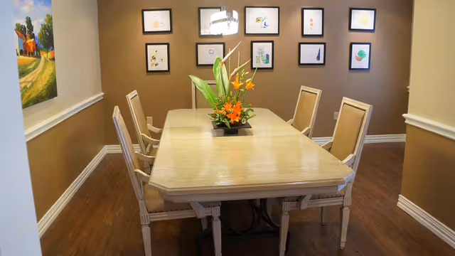 A dining room with a rectangular wooden table surrounded by six chairs. A floral centerpiece with orange flowers and green leaves is placed on the table. The walls are painted brown and beige, decorated with framed artwork and a colorful painting on the left wall. The floor is wooden.