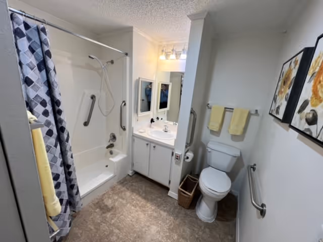 A clean and accessible bathroom featuring a bathtub with a shower and grab bars, a white vanity with a sink and mirror above it, a toilet with grab bars on both sides, two yellow towels hanging on a towel rack, a small wicker basket on the floor, and two framed floral artworks on the wall.
