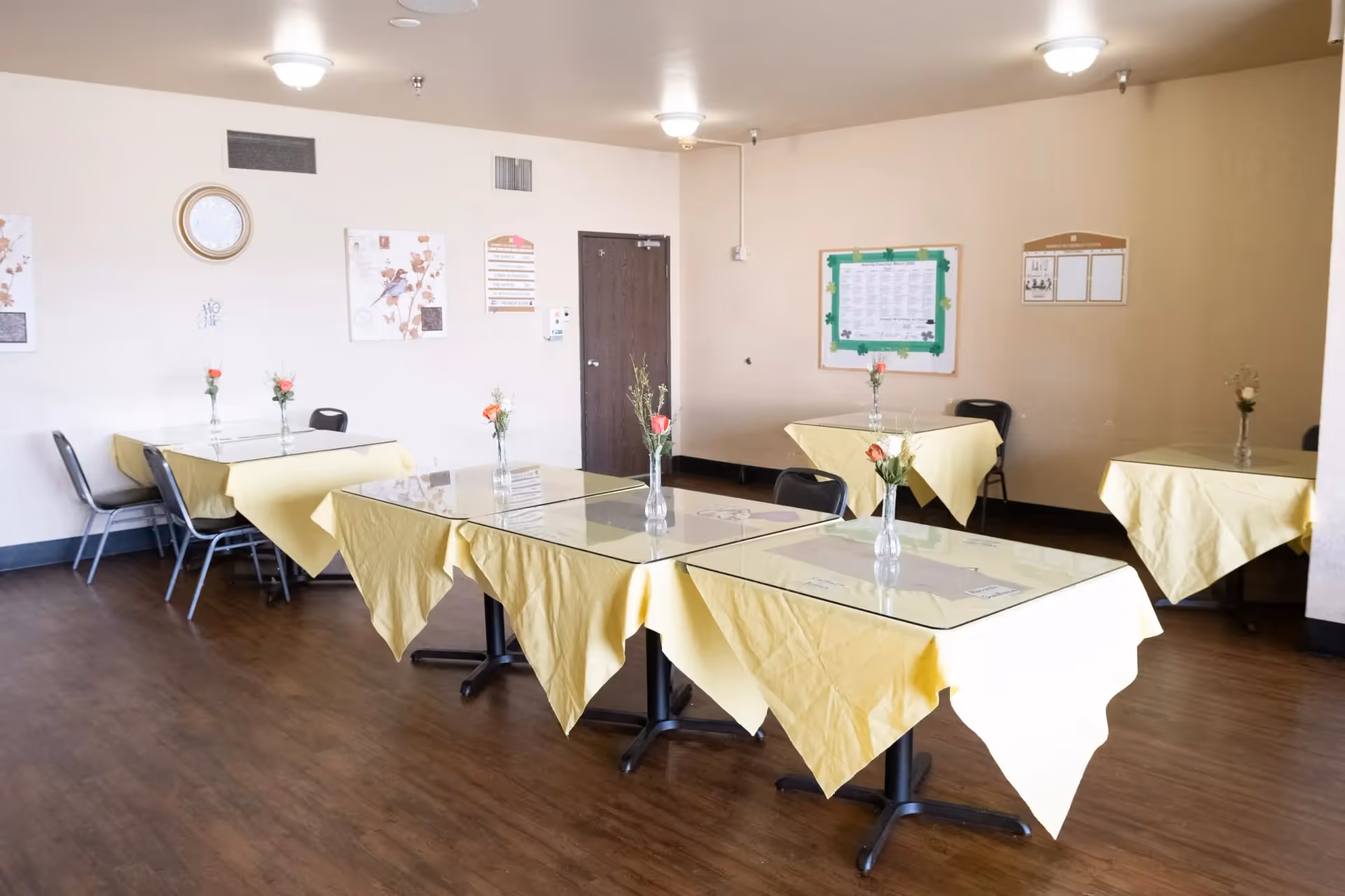 A dining room with several tables covered with yellow tablecloths and glass tops. Each table has a small vase with flowers. The room has wooden flooring, beige walls with some decorations and notices, and several chairs around the tables.