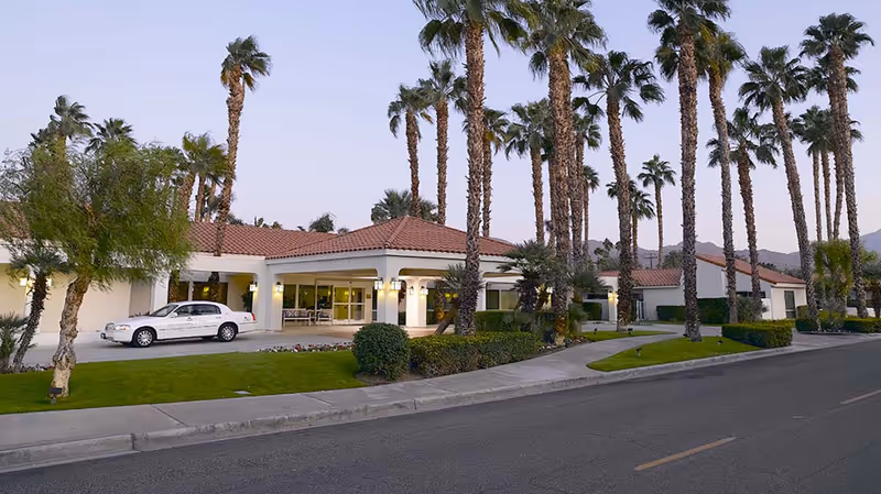 Exterior view of a senior living facility with a tiled roof, surrounded by tall palm trees and well-maintained landscaping. A white car is parked near the entrance, and the sky is clear with a hint of dusk.