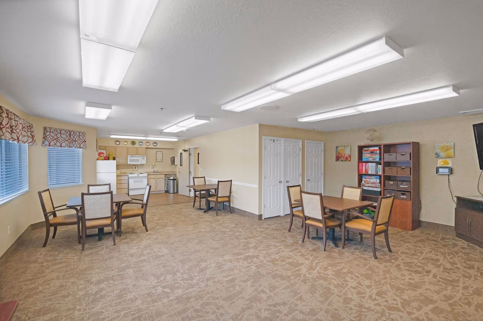 A spacious common area with multiple tables and chairs, a small kitchen with white appliances including a refrigerator, stove, and microwave, and a bookshelf filled with board games and storage bins. The room has beige walls, patterned carpet, and fluorescent ceiling lights. Windows with patterned valances allow natural light in.