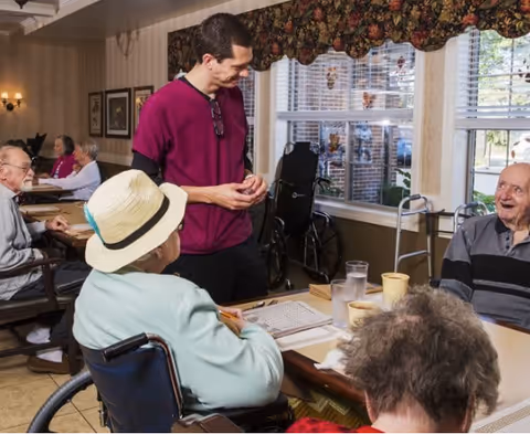 A group of elderly individuals seated around a table in a well-lit room with large windows. A caregiver in a maroon uniform is standing and interacting with them. One elderly person is in a wheelchair wearing a hat, and others are engaged in conversation or activities. The room has floral curtains and framed pictures on the wall.