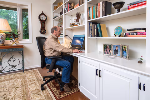 An elderly man sitting at a desk in a home office or study area, working on a laptop. The room features built-in white shelves filled with books, framed photos, and decorative items. A window with a view of greenery and a table lamp are visible on the left side. The floor has a patterned rug, and a wall clock is mounted above the desk.