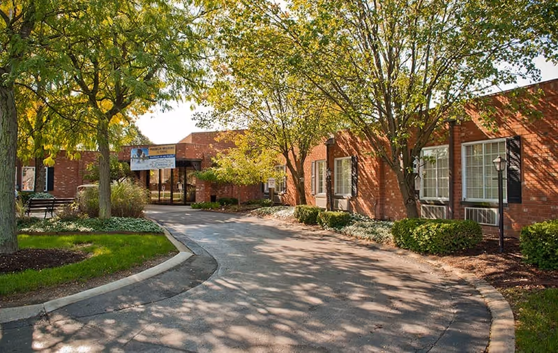 Exterior view of Franklin Meadows facility showing a curved driveway leading to the entrance of a single-story brick building surrounded by trees and landscaped bushes under a sunny sky.