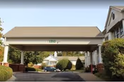 Covered porte-cochere entrance to a senior living building with white columns, a driveway, landscaping, and a parked car.