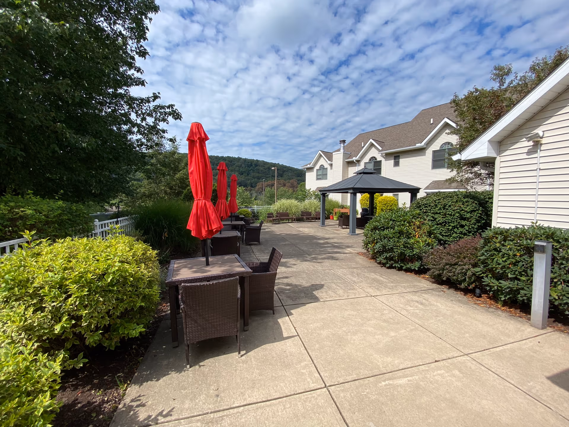 Outdoor patio area at Willowbrook Place with several tables and chairs, each table shaded by a closed red umbrella. The patio is surrounded by green bushes and trees, with a building visible on the right side and a gazebo structure further down the patio. The sky is partly cloudy.