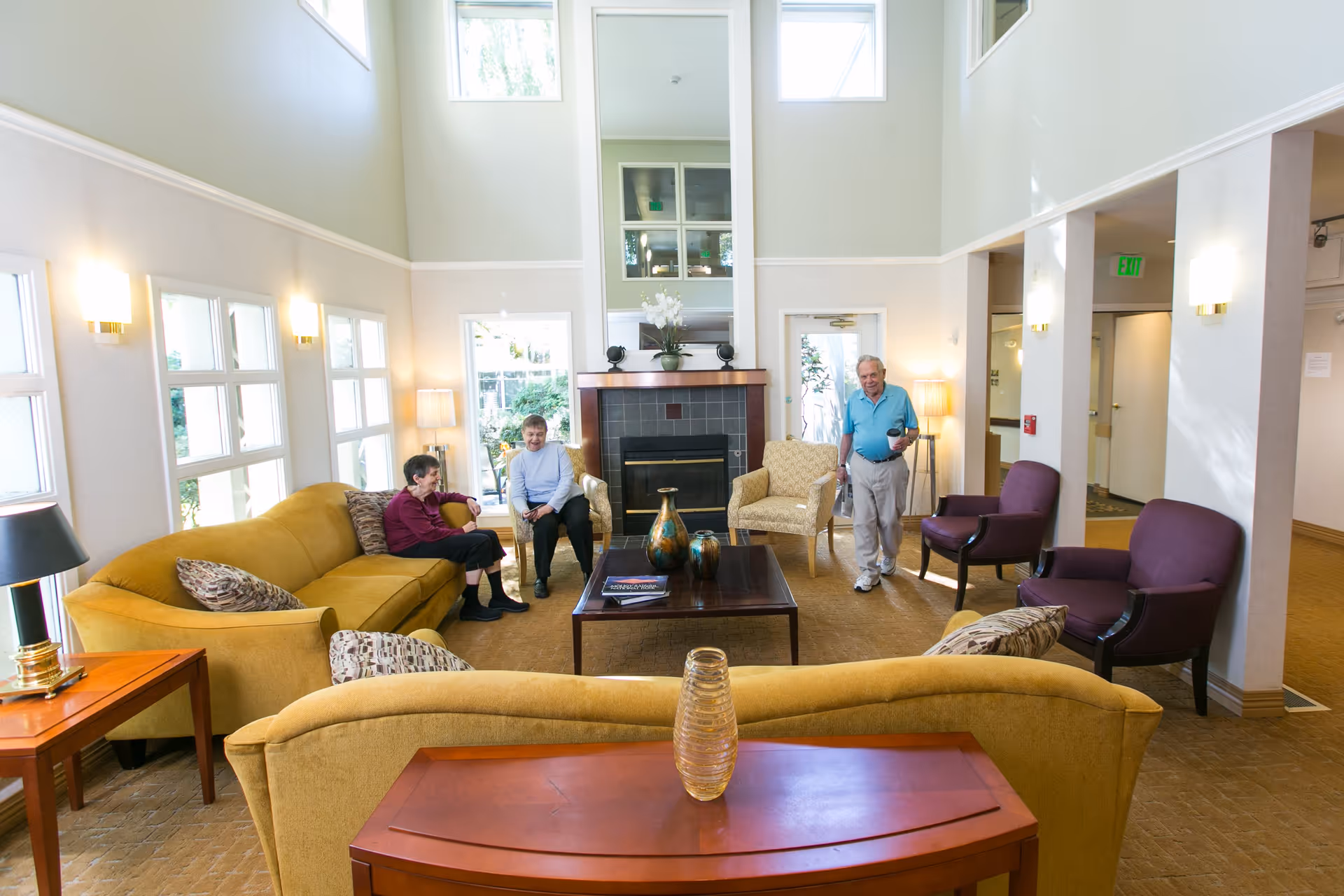 A bright and spacious living room area in Ida Culver House Ravenna with high ceilings, large windows, and a fireplace. The room is furnished with yellow sofas, purple armchairs, and a wooden coffee table with decorative vases. Two elderly women are seated on the sofas, and an elderly man is standing nearby holding a cup.