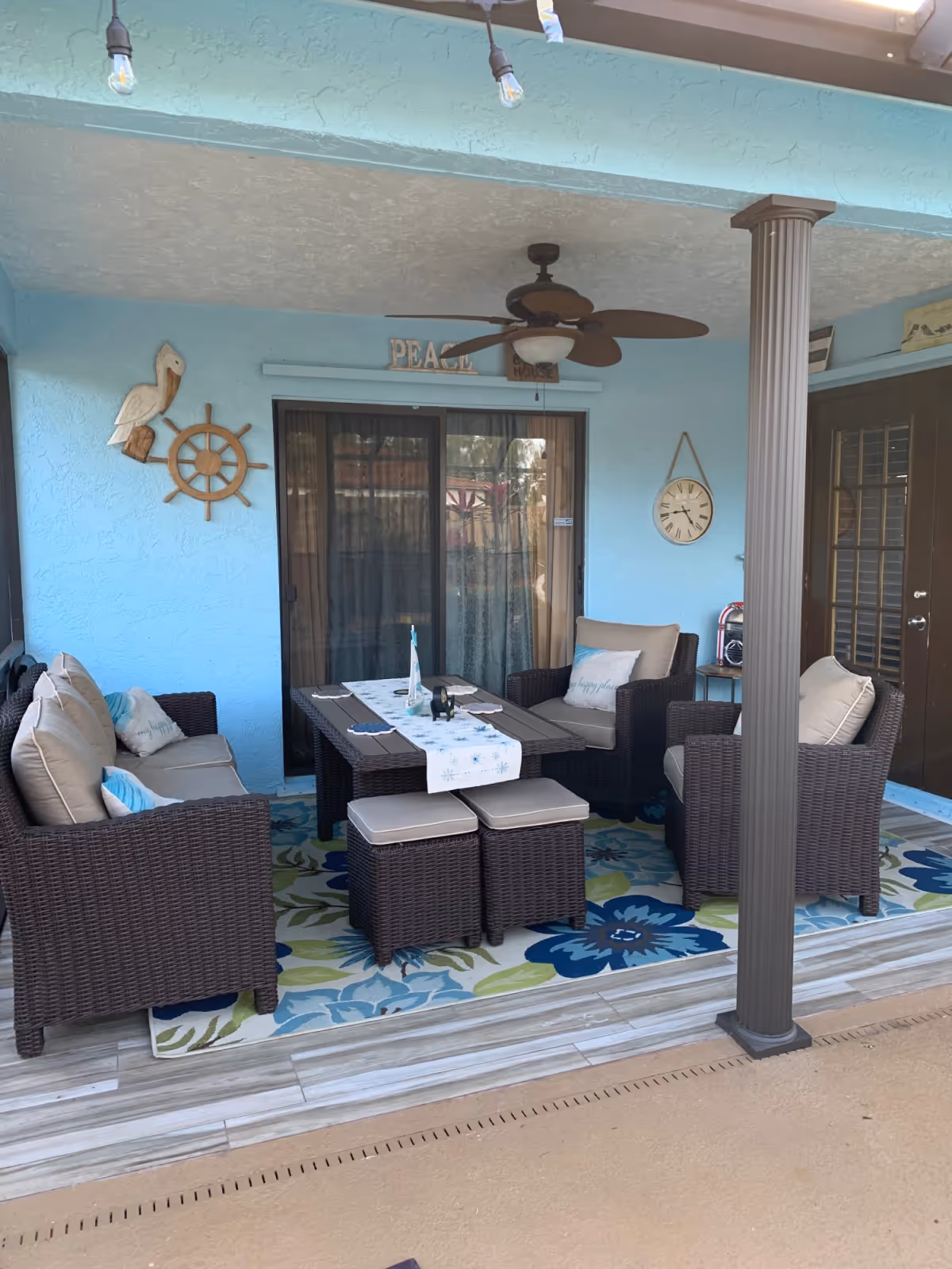 Covered outdoor patio area with blue walls, featuring a ceiling fan, a clock on the wall, and wicker furniture including a sofa, two armchairs, a table with a runner, and two cushioned stools on a floral rug.