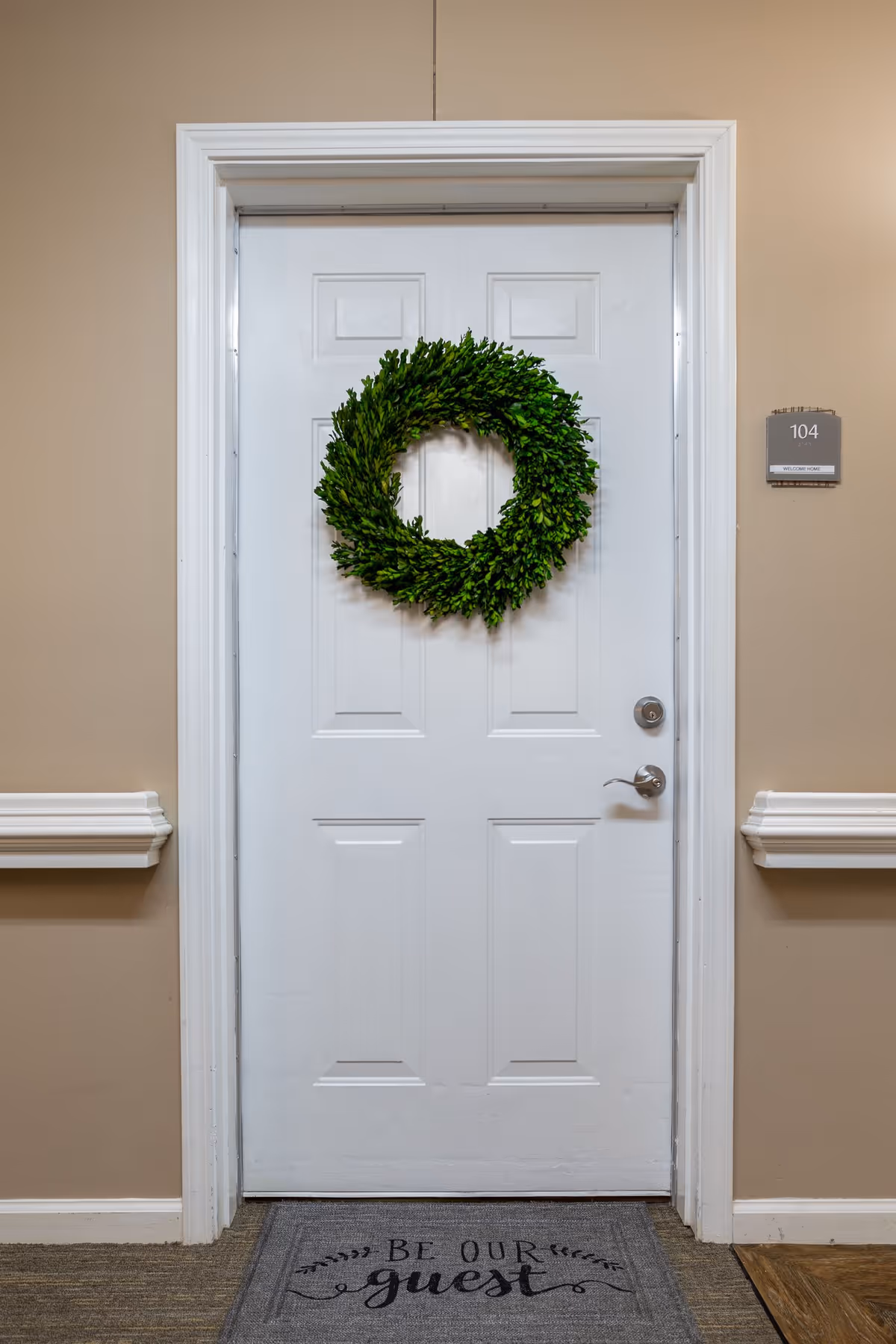A white door with a green wreath hanging in the center. The door has a silver handle and a lock. To the right of the door is a sign with the number 104. On the floor in front of the door is a gray doormat with the words 'Be Our Guest' written on it. The walls around the door are beige with white trim and chair rails.