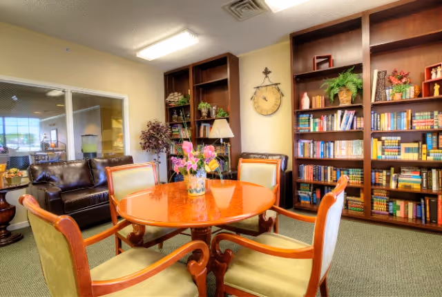 Cozy senior living common room with a round wooden table and chairs, bookshelves, and a leather sofa.