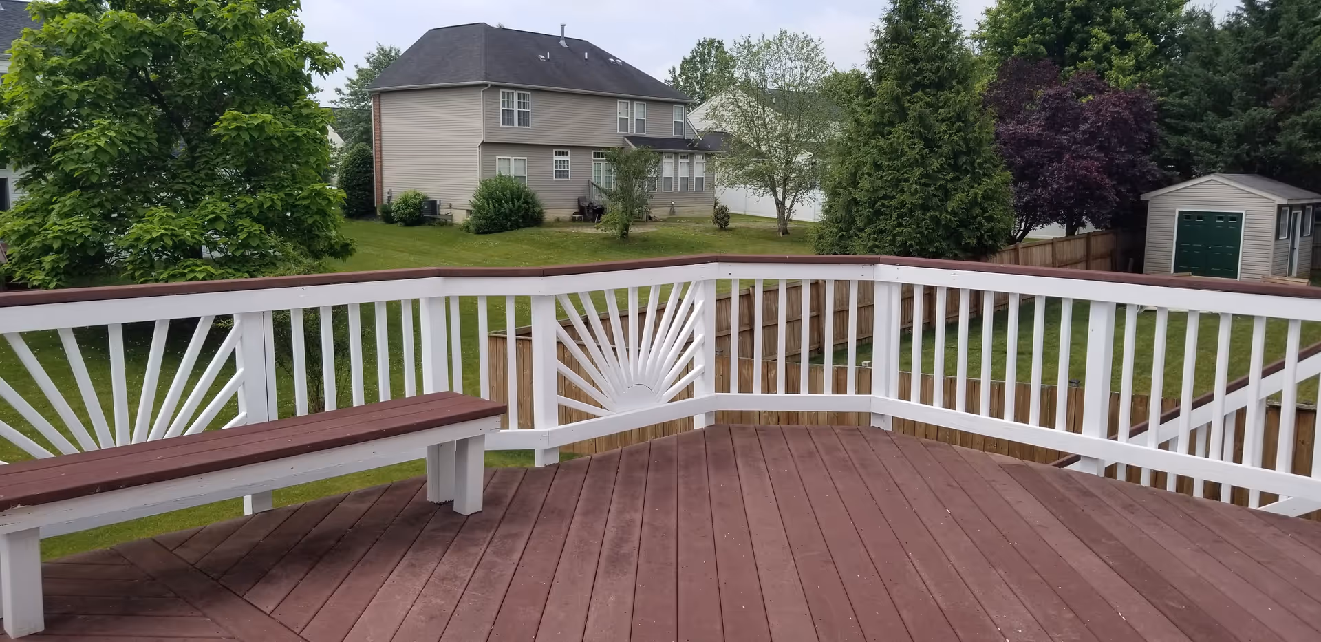View from a wooden deck with white railings and built-in benches overlooking a green backyard with trees, a wooden fence, a shed, and neighboring houses under a cloudy sky.