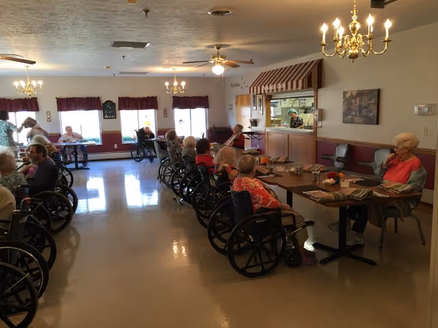 Elderly residents in wheelchairs seated around tables in a communal dining room of a senior living facility.