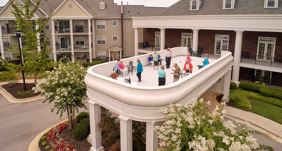 A group of elderly people socializing on a large elevated outdoor terrace with white railings, surrounded by greenery and flowers. The terrace is part of a senior living facility with multi-story buildings in the background.