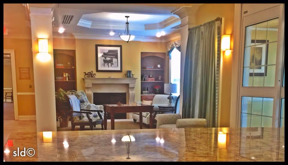 A cozy senior living common area featuring a fireplace with a framed piano picture above it, built-in shelves on either side, and comfortable seating including armchairs and a small table. The room is softly lit with wall sconces and a ceiling light fixture, with large windows covered by curtains allowing natural light in. The foreground shows a polished marble countertop reflecting the room's lighting.