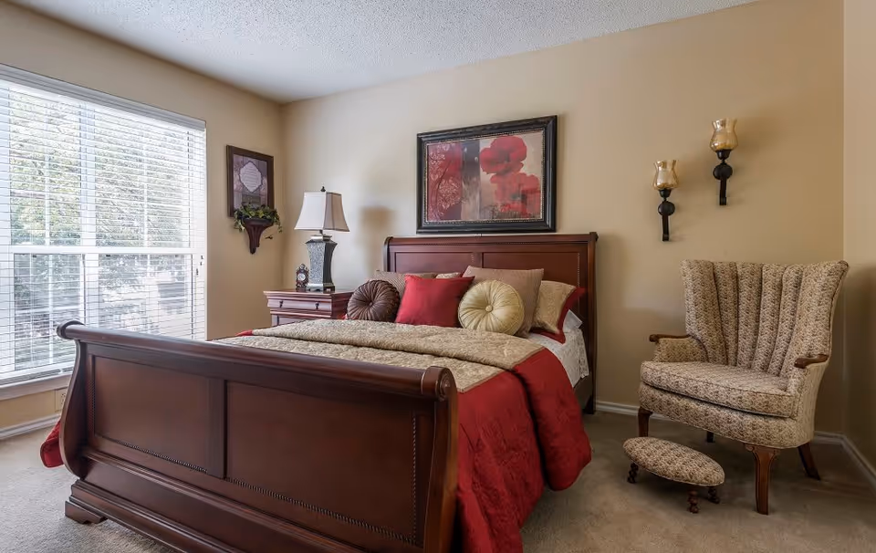 A cozy bedroom with a wooden sleigh bed dressed in red and beige bedding with multiple decorative pillows. There is a wooden nightstand with a lamp and a small clock beside the bed. A framed floral artwork hangs above the bed. To the right, there is a patterned upholstered armchair with a matching footstool. The room has beige walls and a large window with white blinds letting in natural light.