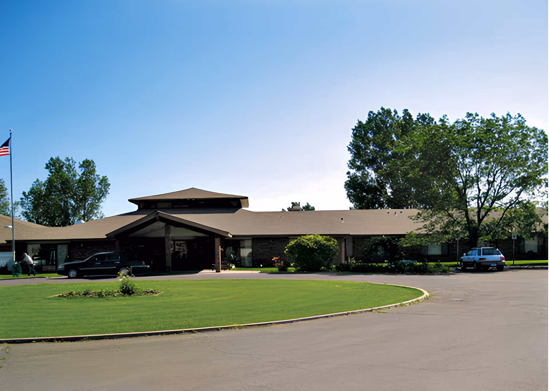 Exterior view of Devonshire Care Center building with a circular driveway, green lawn, trees, and an American flag on a flagpole under a clear blue sky.