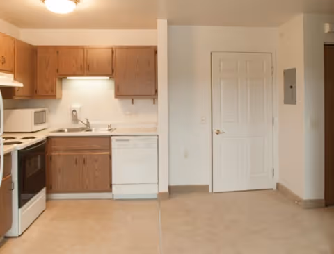 Small kitchenette with oak cabinets, white appliances (stove, microwave, dishwasher), a sink, and a white interior door in a neutral-toned apartment.