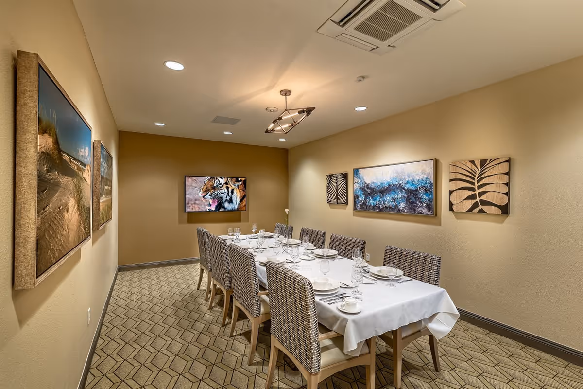 A dining room with a long table set for a meal with white tablecloth, plates, glasses, and silverware. The room has beige walls decorated with framed artwork, including a large painting of a tiger on the far wall. There are ten woven chairs around the table, and the ceiling has recessed lighting and a modern light fixture.