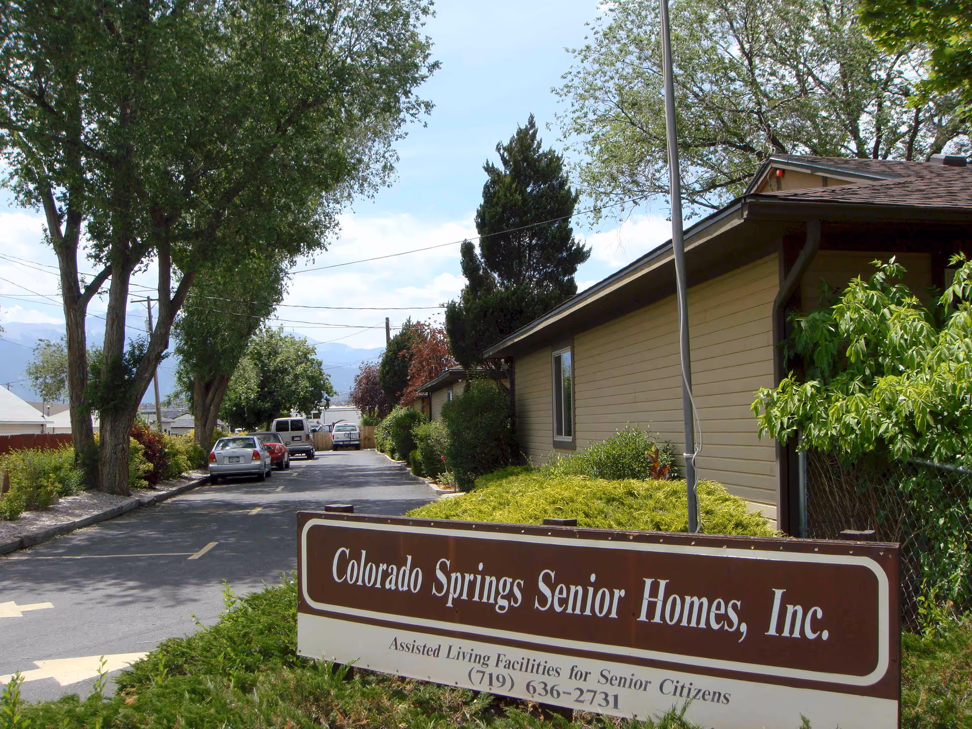 Exterior view of Colorado Springs Senior Homes, Inc. showing a driveway lined with trees and parked cars, with a beige building on the right and a sign in the foreground indicating it is an assisted living facility for senior citizens.