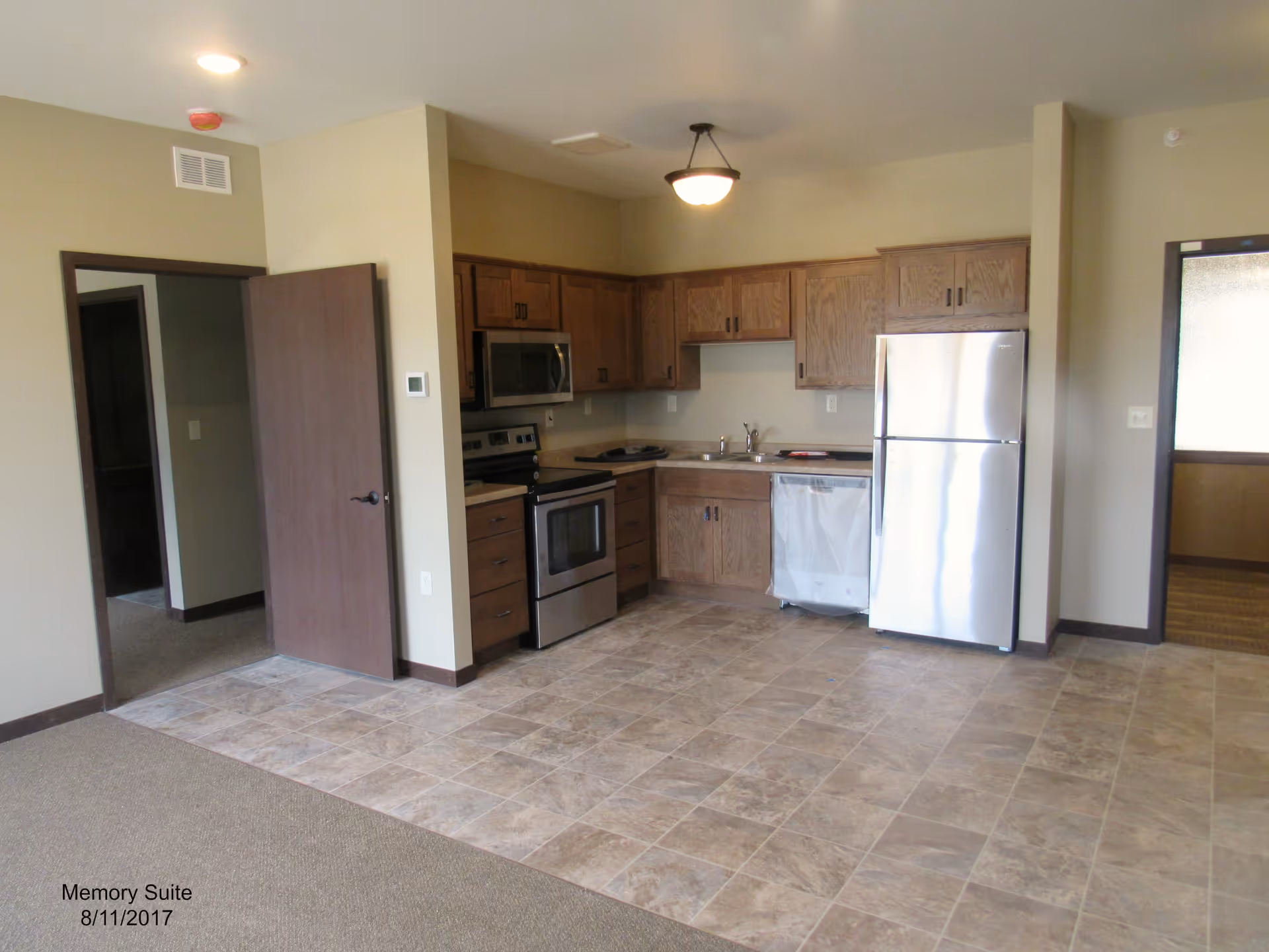 Interior view of a kitchen area in a senior living facility suite. The kitchen features wooden cabinets, a stainless steel refrigerator, stove, microwave, and dishwasher. The floor is tiled in the kitchen area and carpeted in the adjacent room. There are two open doorways leading to other rooms.