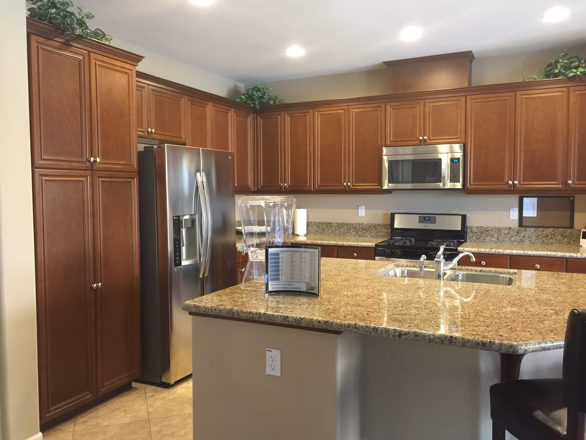 Modern kitchen with wooden cabinets, stainless steel refrigerator, microwave, and stove. A granite countertop island with a sink and faucet is in the foreground, along with a clear water dispenser and a small sign on the counter. The kitchen is well-lit with recessed ceiling lights and has decorative greenery on top of the cabinets.