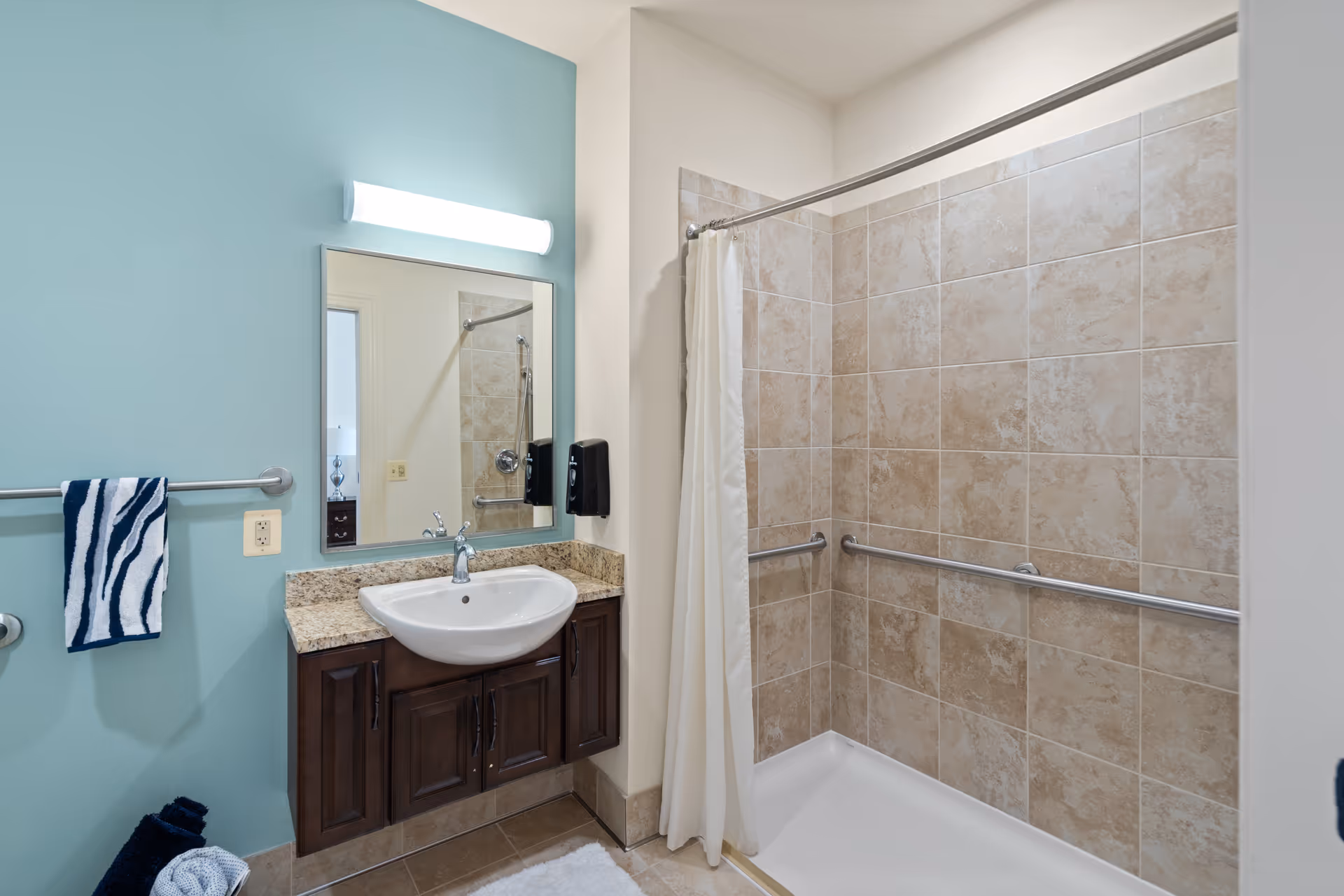 A clean bathroom featuring a tiled walk-in shower with grab bars and a white shower curtain. There is a sink with a granite countertop and dark wooden cabinets below. Above the sink is a rectangular mirror with a light fixture. A towel rack on the left wall holds a blue and white striped towel.